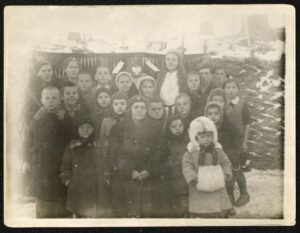 Na zdjęciu widać grupę dzieci stojących na tle dekoracji z orłem w koronie. Przed nimi siedzi kobieta w mundurze. Wokół widać śnieg.