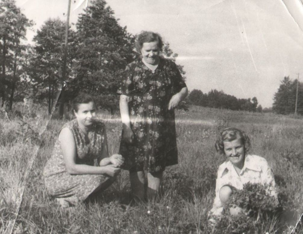Na tym czarnobiałym zdjęciu widzimy trzy kobiety. Dwie kucają, jedna trzyma w rękach bukiet polnych kwiatów.