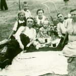 Na czarno-białym zdjęciu widać rodzinę leżącą na kocu. Na kocu leżą też produkty spożywcze, jest to piknik.