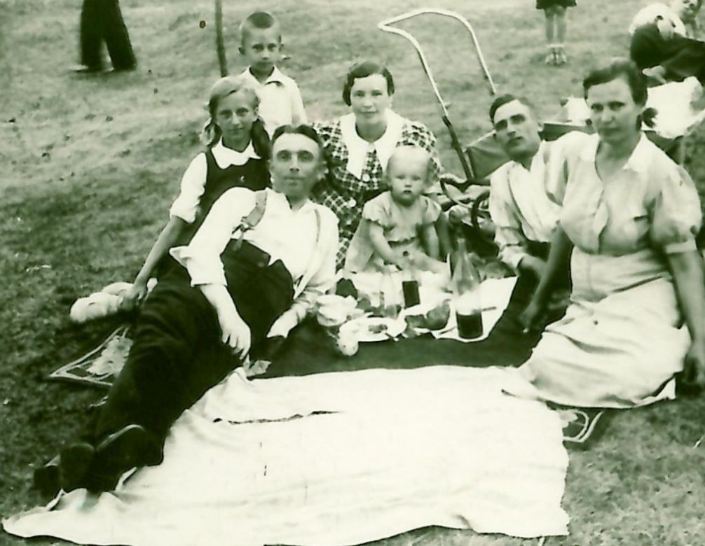 Na czarno-białym zdjęciu widać rodzinę leżącą na kocu. Na kocu leżą też produkty spożywcze, jest to piknik.