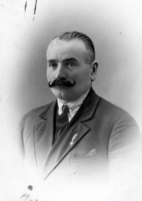Zdjęcie przedstawia mężczyznę z wąsami w marynarce, koszuli i krawacie