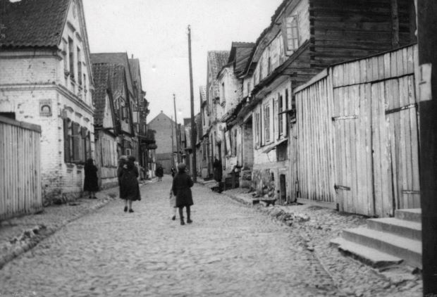 Na czarno-białym zdjęciu widać ludzi idących brukowaną ulicą zabudowaną z obu stron domami.