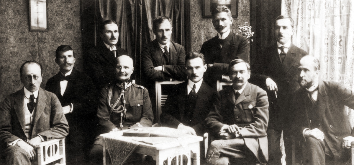Na zdjęciu widac grupę 10 mężczyzn w eleganckich strojach w pomieszczeniu. 4 z nich stoi, pozostali siedzą.