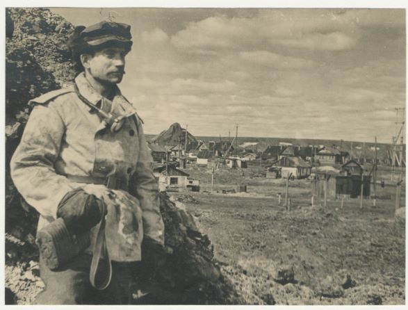 Na zdjęciu widać mężczyznę z wąsami, stojącego na tle osady położonej w górach