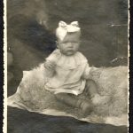 Fotografia małej dziewczynki z kokarda na głowie, siedzącej na owczej skórze