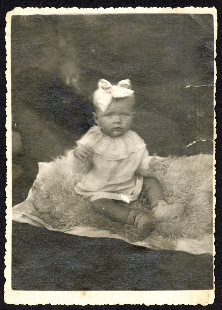 Fotografia małej dziewczynki z kokarda na głowie, siedzącej na owczej skórze