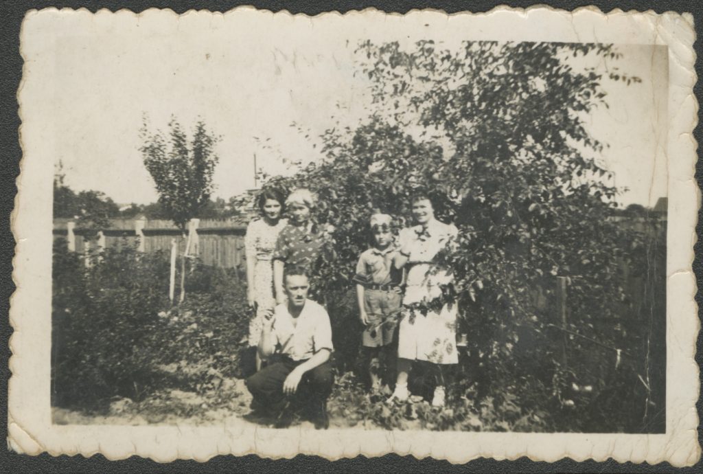 Na zdjęciu widać pięć osób w ogrodzie, trzy stojące kobiety, kucającego mężczyznę i chłopca w harcerskim mundurze