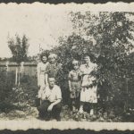 Na zdjęciu widać pięć osób w ogrodzie, trzy stojące kobiety, kucającego mężczyznę i chłopca w harcerskim mundurze