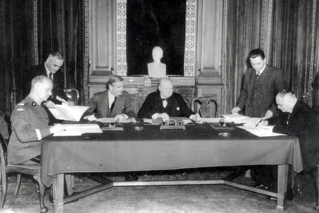 Na zdjęciu widać czterech mężczyzn siedzących przy stole i dwóch stojących po jego obu stronach.