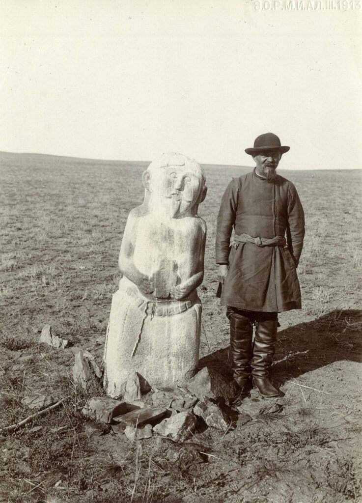 Na zdjęciu widać mężczyznę w kapeluszu stojącego obok kamiennej rzeźby wyobrażającej człowieka.