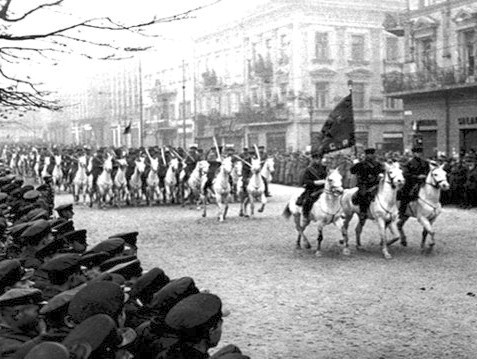 Na zdjęciu widać wojsko defilujące na koniach wśród miejskich zabudowań.