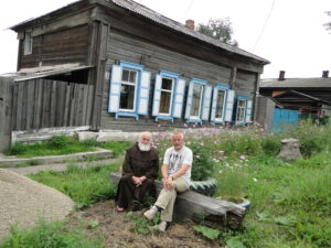 Dwóch mężczyzn (jeden z nich w habicie) siedzących przed drewnianym budynkiem