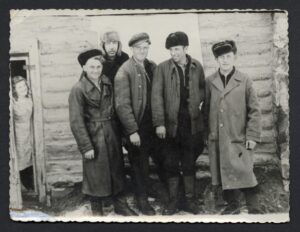 A group of men in coats standing by the  wooden wall of the house