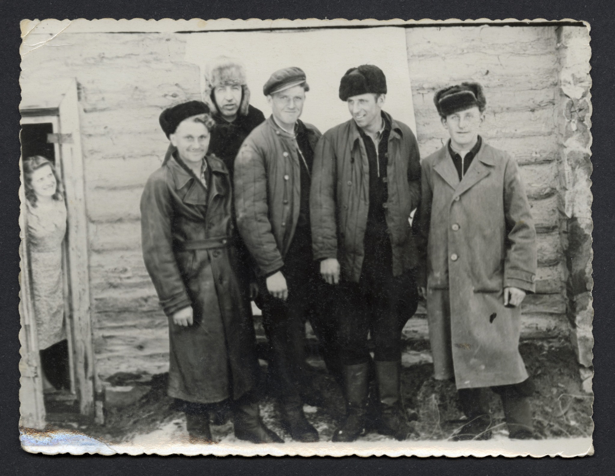 Grupa mężczyzn w płaszczach, kurtkach i czapkach stojących na tle ściany drewnianego budynku