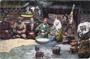Grupa kobiet, mężczyzn i dzieci w orientalnych strojach, siedzących wewnątrz pomieszczenia. Kazachowie