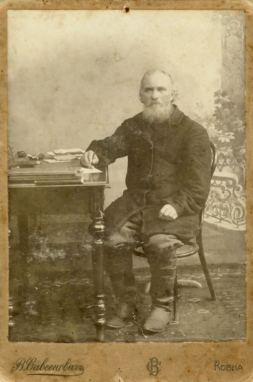 Portret fotograficzny w atelier przedstawiający starszego mężczyznę z broda siedzącego przy stole i piszącego na kartce papieru.