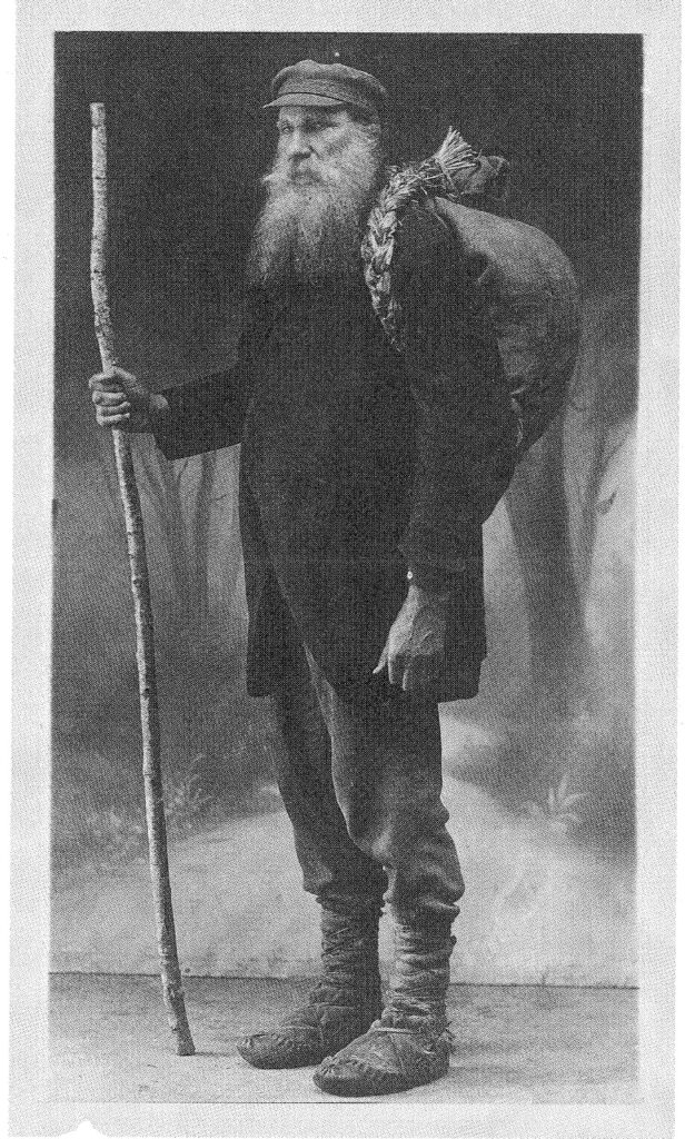 Portret fotograficzny w atelier przedstawiający stojącego mężczyznę z brodą w czapce, ciepłym ubraniu, worku na plecach i kijem w dłoni.