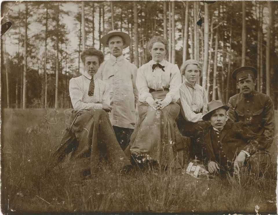 Fotografia przedstawiająca trzy kobiety i trzech mężczyzn siedzących na trawie na tle ściany lasu. Jeden mężczyzna w mundurze, dwaj w kapeluszach i surdutach, kobiety w spódnicach i koszulach.
